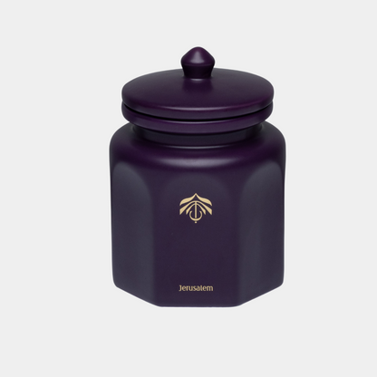 A dark purple hexagonal ceramic candle vessel with a matching lid and gold logo, set against a plain light gray background.