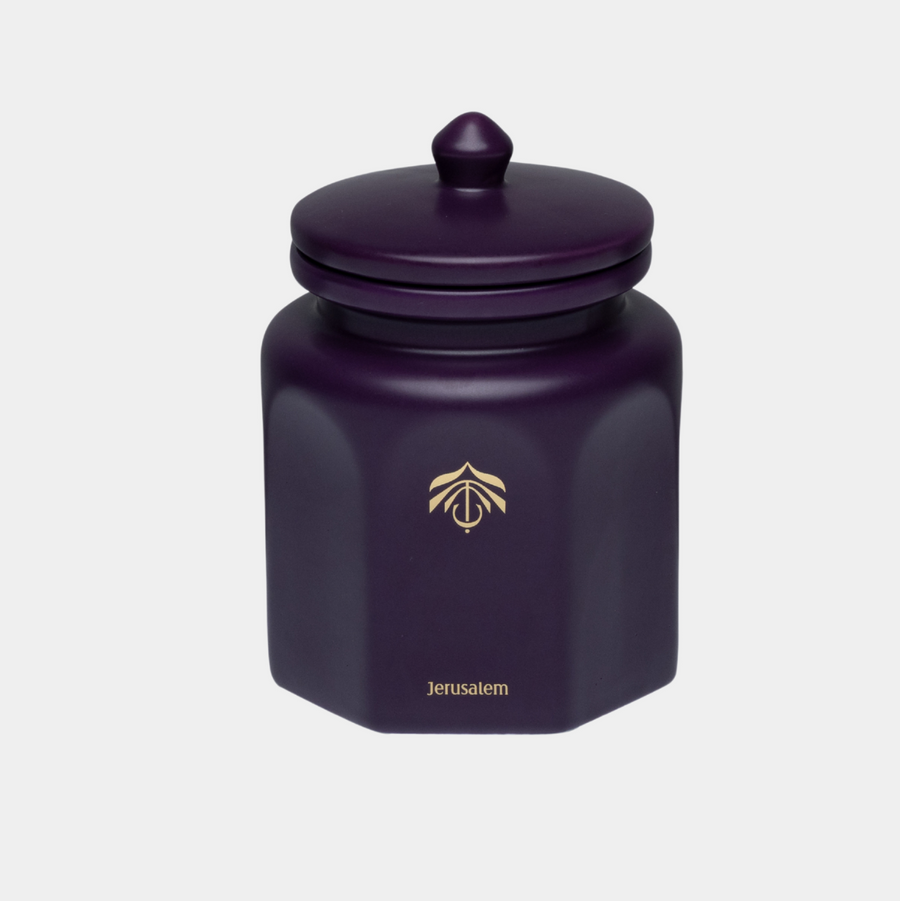 A dark purple hexagonal ceramic candle vessel with a matching lid and gold logo, set against a plain light gray background.