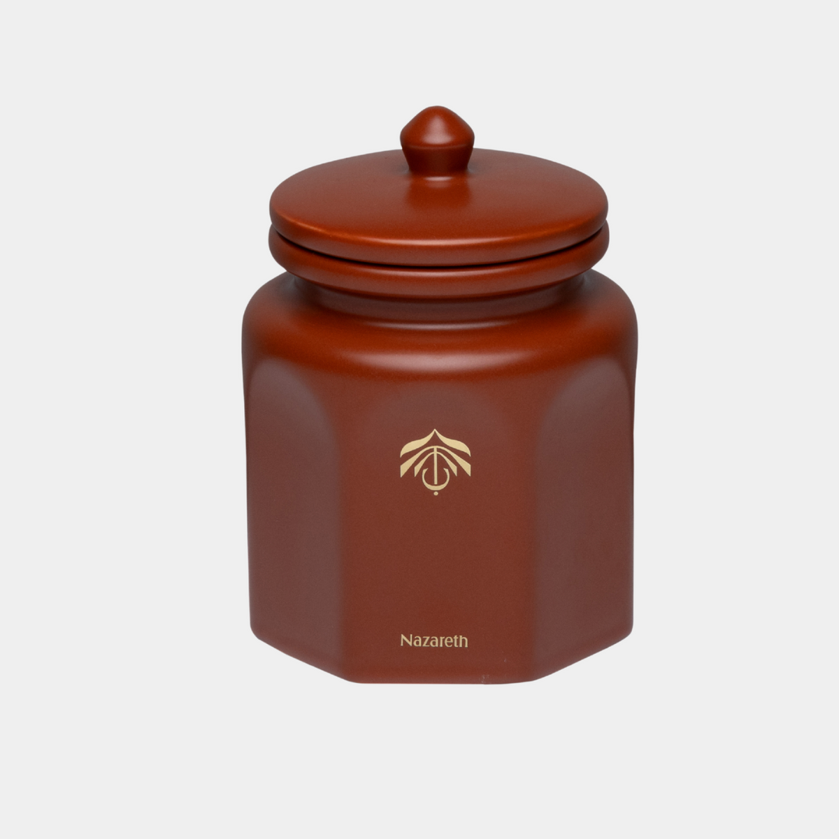 A terracotta brown hexagonal ceramic candle vessel with a matching lid, featuring a gold logo and the candle name 'Nazareth' printed on the front, set against a plain light gray background.