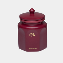 A dark red hexagonal candle vessel with a matching lid, featuring a gold logo and the text 'Garden of Eden' on the front, set against a light gray background.
