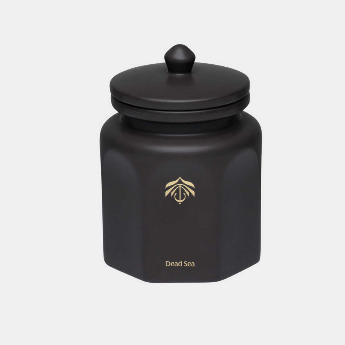 A matte black hexagonal cande vessel with a matching lid and a small gold logo and 'Dead Sea' text on the front, set against a light gray background.