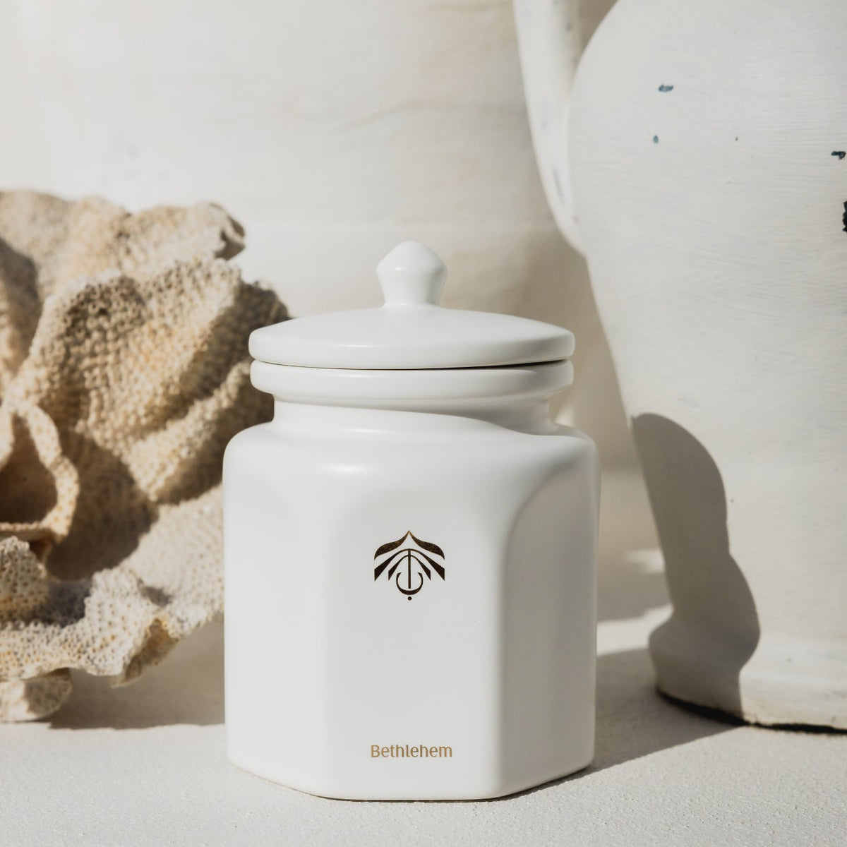 A white ceramic candle vessel with a matching lid, featuring a gold logo and the brand name 'Bethlehem' on the front, styled on a textured white surface with warm natural shadows.