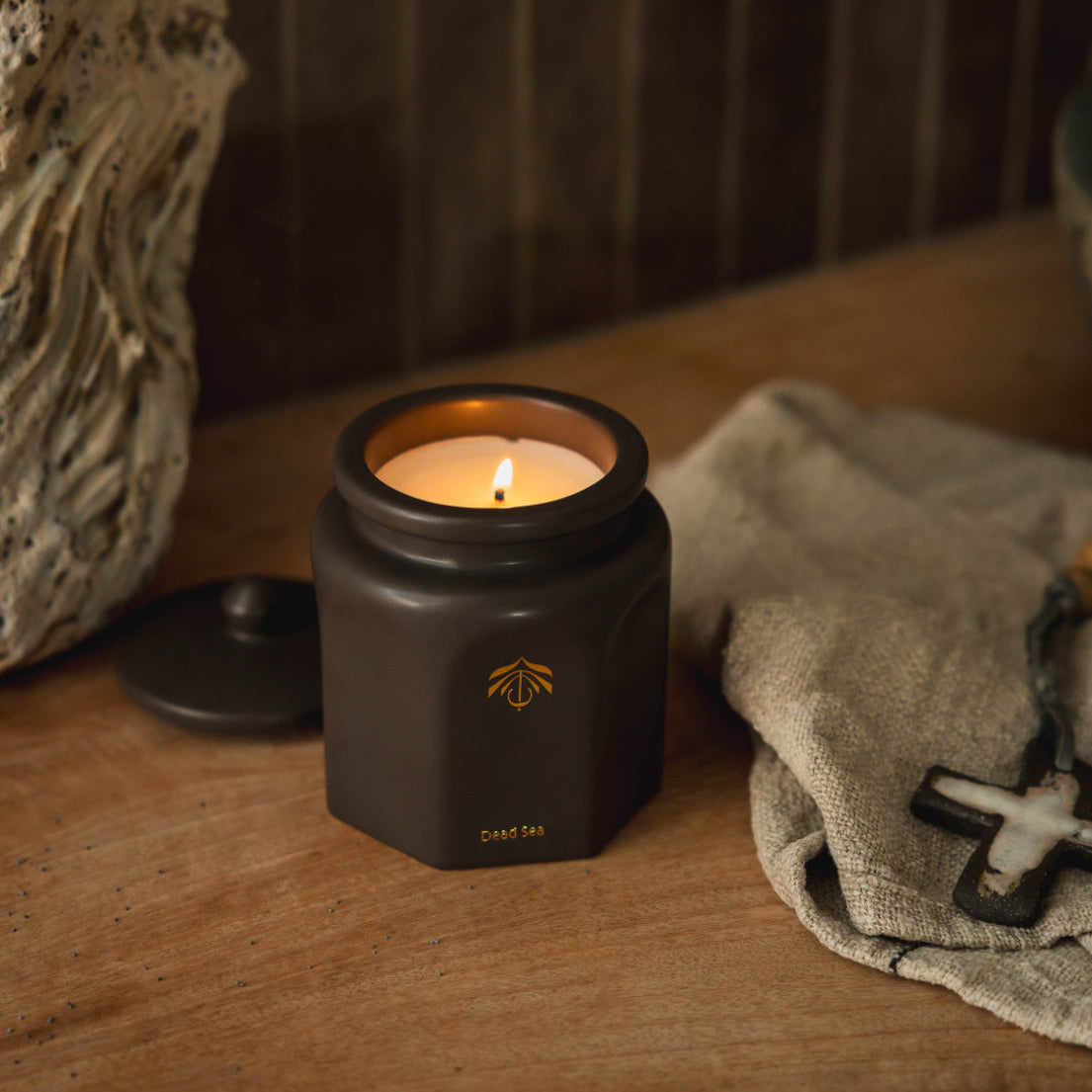 A matte black hexagonal candle vessel with a gold logo and 'Dead Sea' text, lit on a rustic wooden table next to a textured stone and a linen cloth.