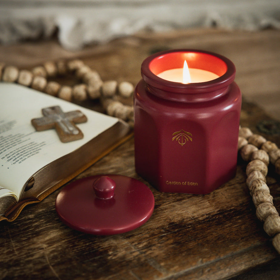 A lit red candle vessel with its lid off, placed on a rustic wooden surface beside an open book and a wooden cross, with a beaded rosary draped around it.