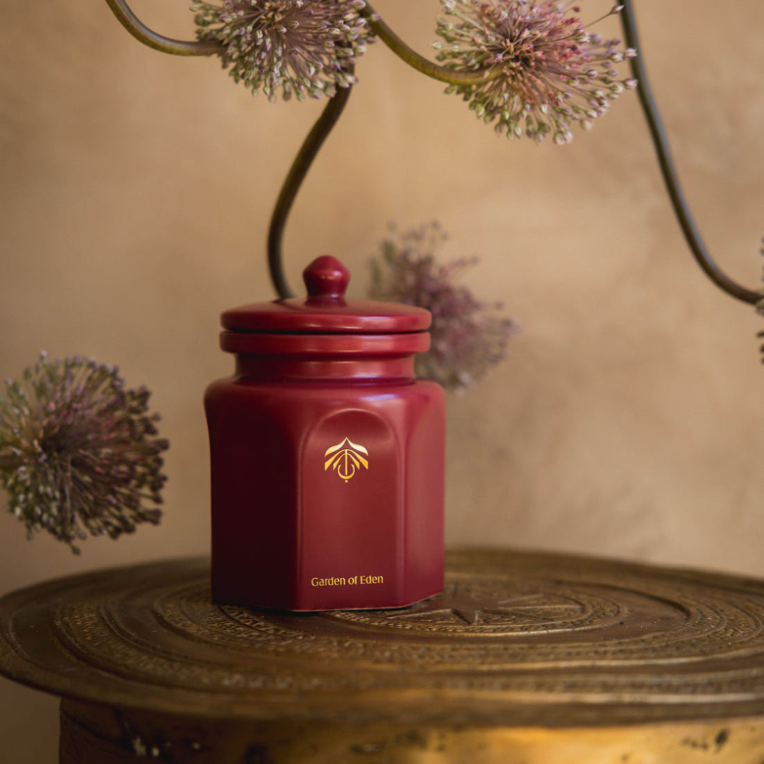 A red candle vessel with a gold logo and the text 'Garden of Eden' on the front, styled on a rustic wooden surface with dried flowers against a warm beige background.