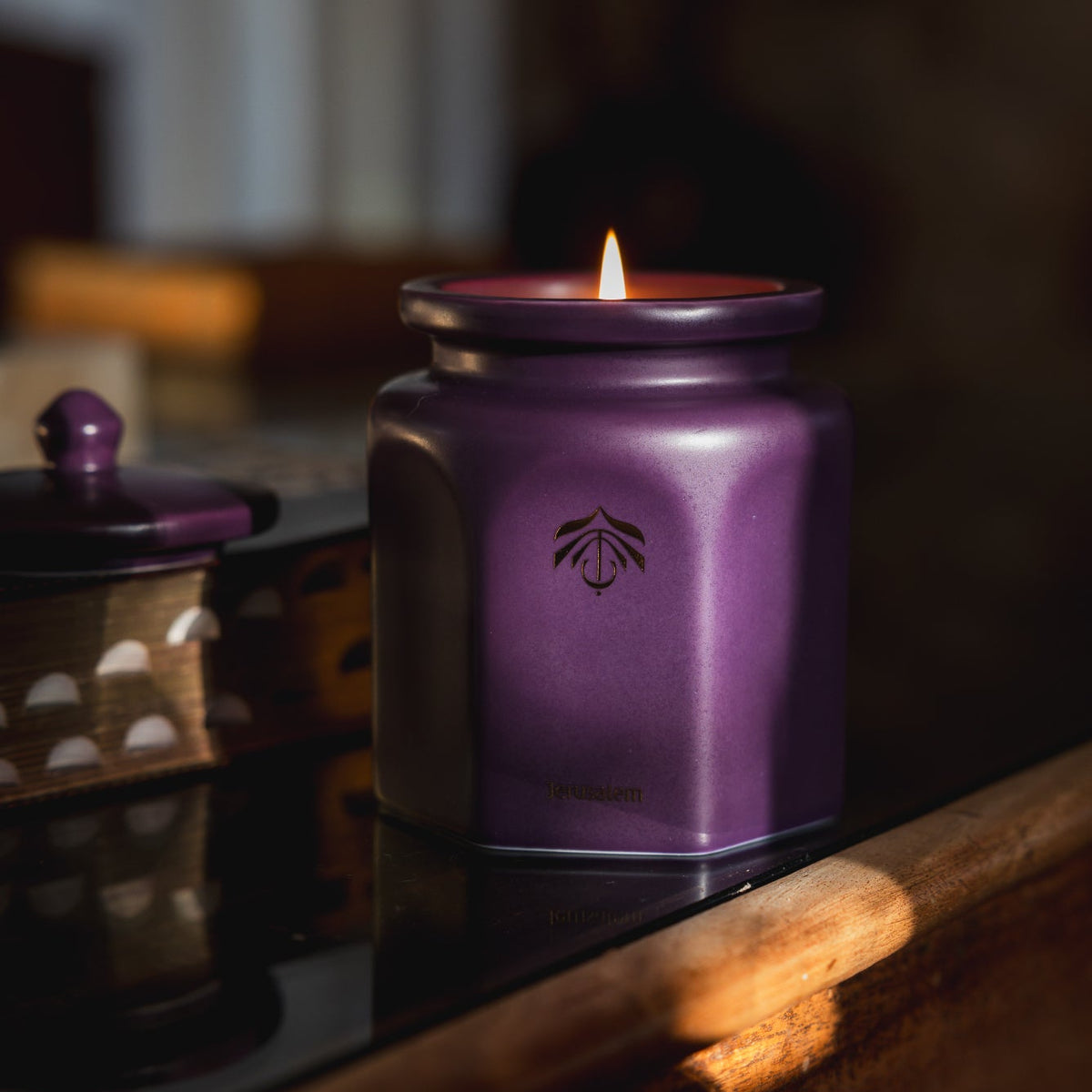 A lit purple hexagonal candle in a matching metal lid sits on a dark reflective surface in a warm, dimly lit interior setting.
