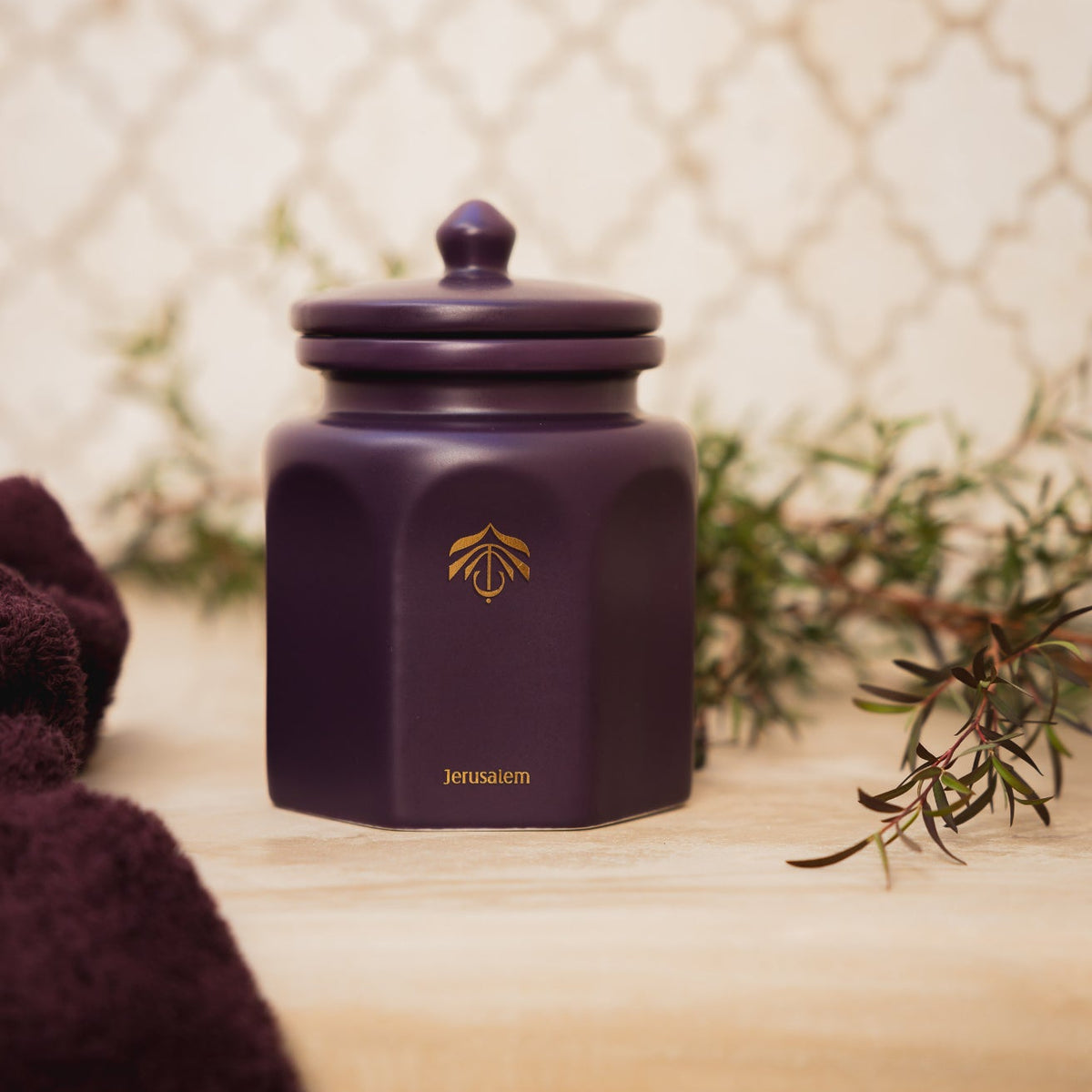 A dark purple hexagonal cnadle vessel with a matching lid, featuring a gold logo and the brand name 'Jerusalem' on the front, styled on a light wooden surface with rosemary and a dark blanket.