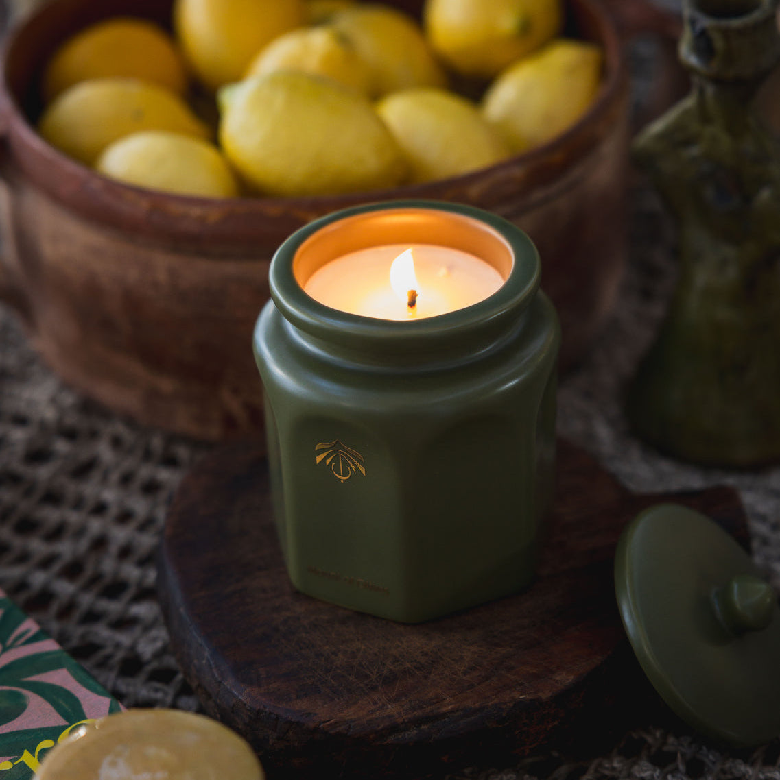 A green hexagonal candle vessel with a gold logo, lit and set on a wooden coaster, with a matching lid nearby, a bowl of lemons in the background.