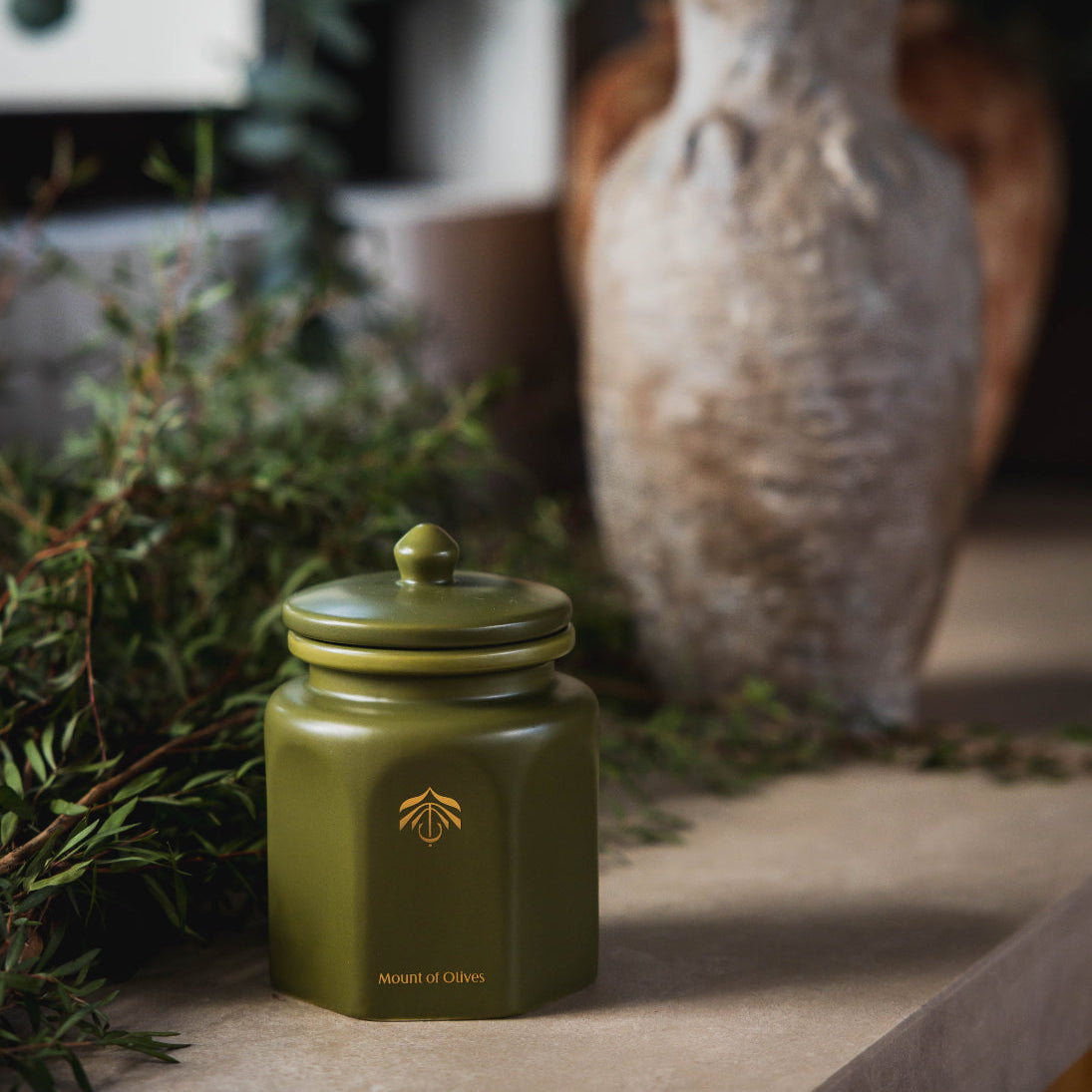 An olive green ceramic candle vessel with a matching lid and a gold logo, styled on a stone surface with greenery and a textured vase in the background.