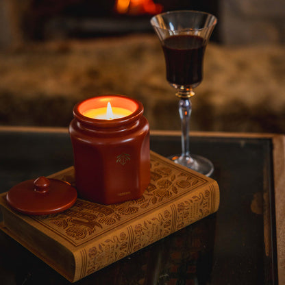 A warm, cozy scene featuring a lit terracotta ceramic candle vessel with its lid resting beside it on a bible, with a glass of dark red wine in front of a blurred fireplace.