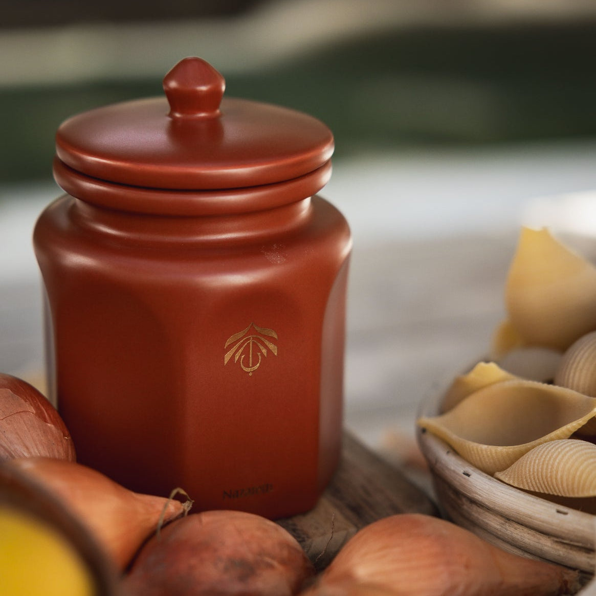 A terracotta-colored ceramic candle vessel with a matching lid, featuring a small gold logo on the front, styled among various onions, garlic, and pasta shells on a rustic wooden surface.