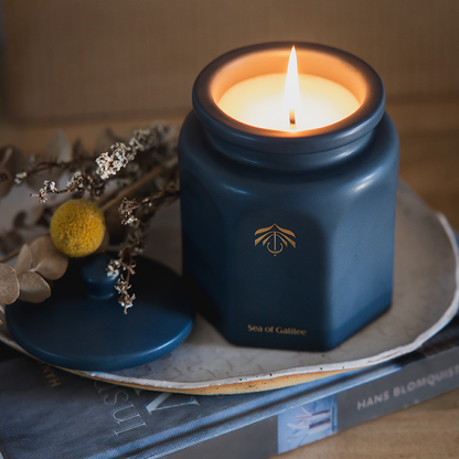 A lit teal blue hexagonal candle in a matching jar sits on a tray atop stacked books, with dried flowers beside it. The candle vessel has a gold logo and the text 'Sea of Galilee'.