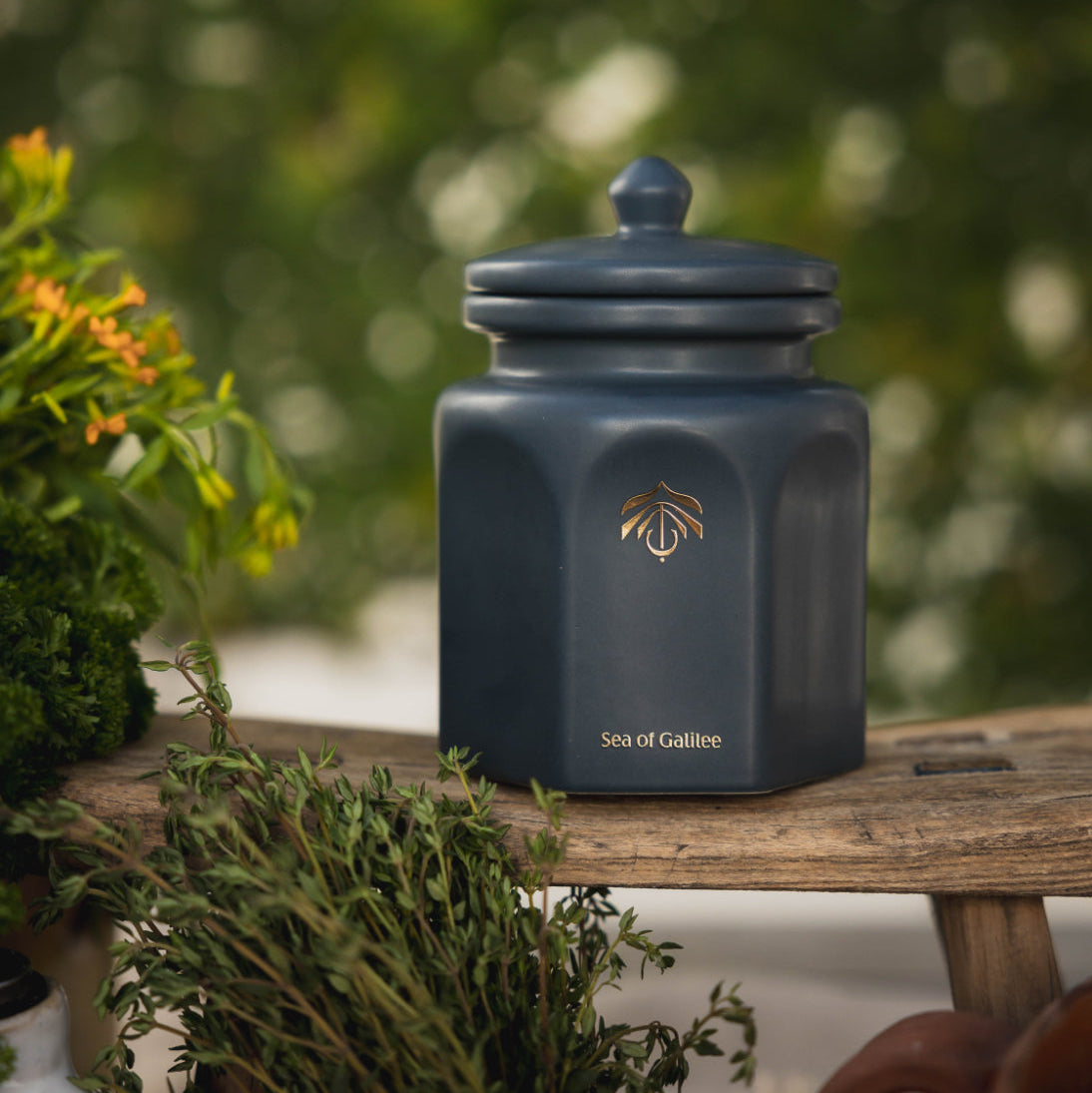 A dark blue-gray ceramic candle vessel with a lid, featuring a gold logo and the text 'Sea of Galilee' on the front, displayed on a rustic wooden bench with green foliage in the background.