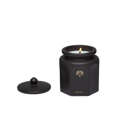 A dark purple hexagonal candle vessel with a matching lid, shown on a plain white background. The candle is lit with a small flame, and a gold logo and the candle name 'Dead Sea' are printed on the front of the jar.