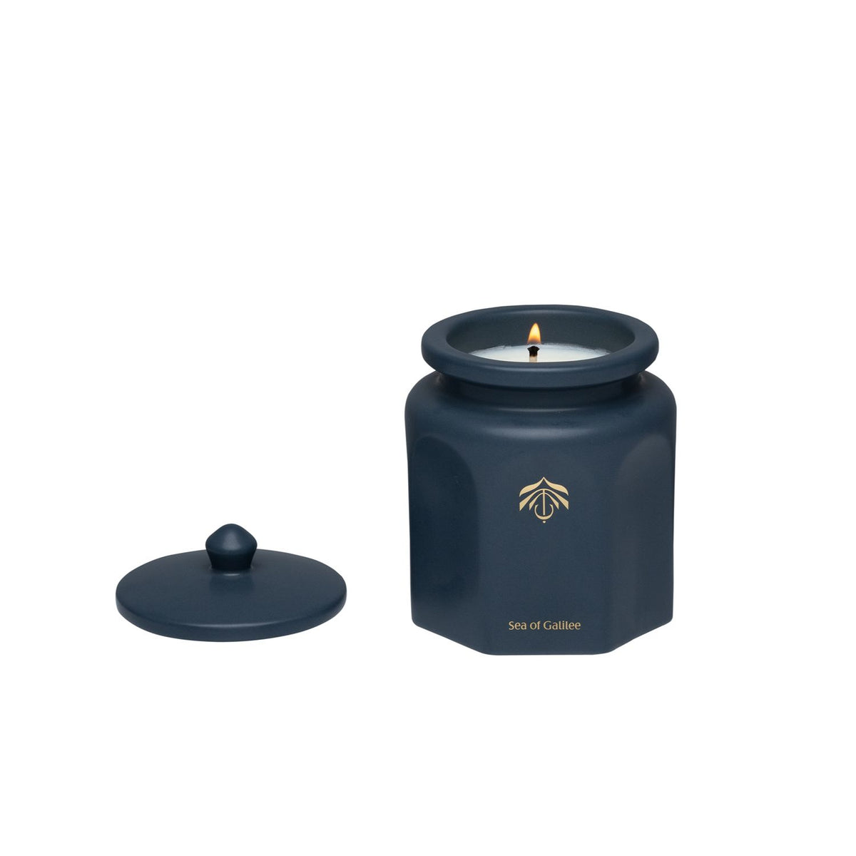 A dark blue hexagonal candle vessel with a matching lid, shown on a plain white background. The candle is lit with a small flame, and a gold logo and the Candle name 'Sea of Galilee' are printed on the front of the jar.