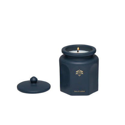 A dark blue hexagonal candle vessel with a matching lid, shown on a plain white background. The candle is lit with a small flame, and a gold logo and the Candle name 'Sea of Galilee' are printed on the front of the jar.